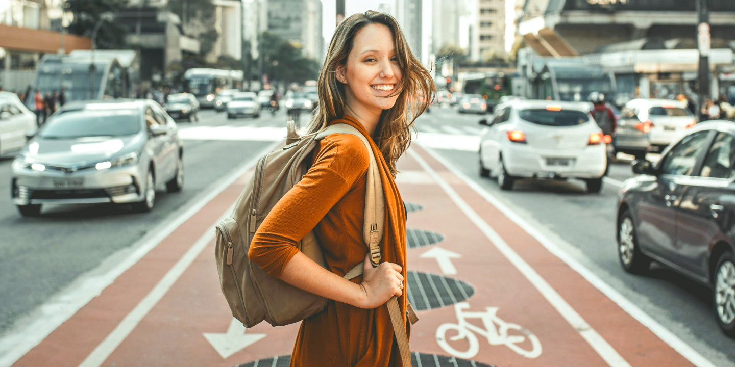 Eine junge Fraue steht auf einem Fahrradstreifen mit ihrem Rucksack und links und rechts fahren die Autors vorbei.