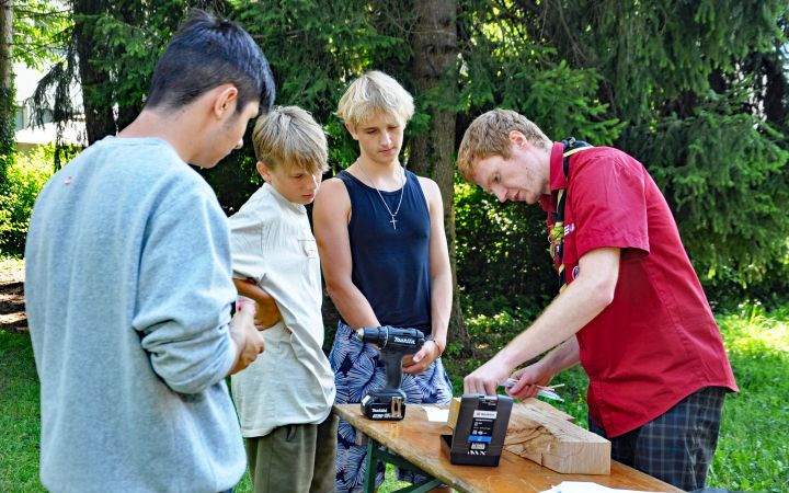 3 Jugendliche arbbeiten an einem Holzstück gemeinsam mit einem Erwachsenen von den Pfadfindern